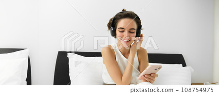 Portrait of smiling young woman in wireless headphones, sitting on bed with mobile phone, listening music in earphones 108754975