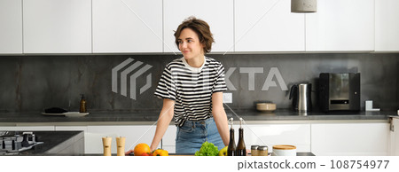 Portrait of smiling cute young woman making breakfast, chopping vegetables in the kitchen, preparing vegan meal 108754977