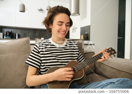 Portrait of young modern woman, musician playing ukulele at home, picking chords for new song, sitting on sofa in living room and smiling 108755006