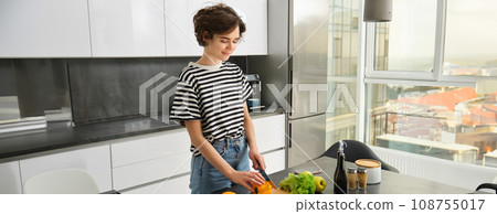 Portrait of smiling cute young woman making breakfast, chopping vegetables in the kitchen, preparing vegan meal Portrait of smiling cute young woman making breakfast, chopping vegetables in the kitchen, preparing vegan meal 108755017
