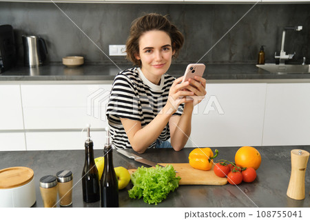 Portrait of modern young woman thinking what to cook, sitting in the kitchen with smartphone and vegetables, making a meal, healthy diet salad Portrait of modern young woman thinking what to cook, sitting in the kitchen with smartphone and vegetables, making a meal, healthy diet salad 108755041