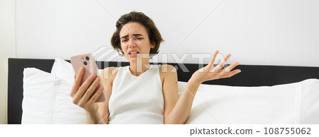 Portrait of confused woman lying in bed, looking at smartphone and shrugging, looking puzzled at mobile screen with frowned face expression 108755062