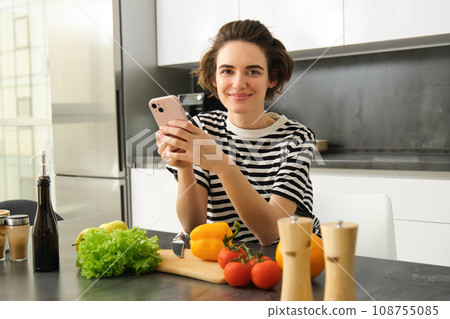 Portrait of young woman standing in the kitchen with vegetables and chopping board, holding smartphone, searching healthy diet recipes on mobile phone browser, cooking at home Portrait of young woman standing in the kitchen with vegetables and chopping board, holding smartphone, searching healthy diet recipes on mobile phone browser, cooking at home 108755085