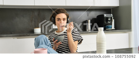 Cute girl eats her breakfast, holding bowl of cereals with milk, drinks coffee and listens to music in wireless headphones, using earphones while having her morning meal 108755092
