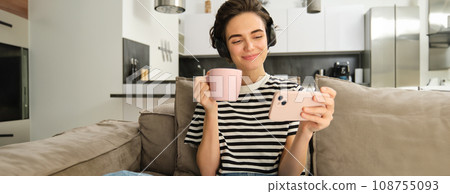Smiling girl watching tv show on smartphone, wearing headphones, drinking tea and sitting on sofa 108755093