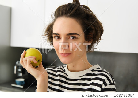 Close up portrait of smiling woman in the kitchen, holding an apple, eating fruit, having a healthy snack for lunch at home 108755094