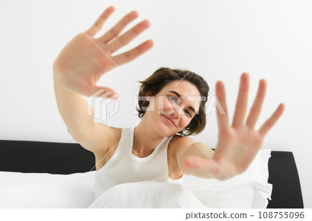 Beautiful young woman stretching her hands towards camera, waking up in morning, resting in her bed, smiling and looking happy 108755096