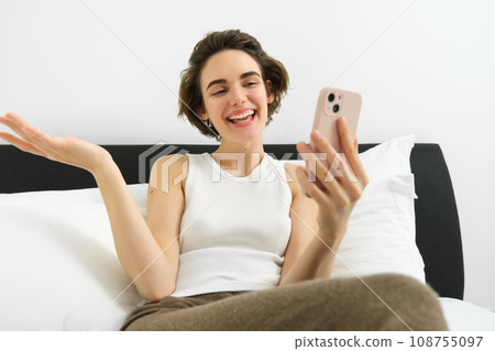 Smiling friendly young woman chatting on smartphone app, talking via video call app on mobile phone, lying in bedroom, resting in bed and communicating with someone 108755097
