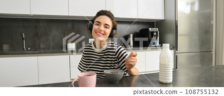 Portrait of happy smiling woman eating cereals with milk, drinking tea and listening music in wireless headphones, enjoys her morning, having breakfast alone in the kitchen Portrait of happy smiling woman eating cereals with milk, drinking tea and listening music in wireless headphones, enjoys her morning, having breakfast alone in the kitchen 108755132