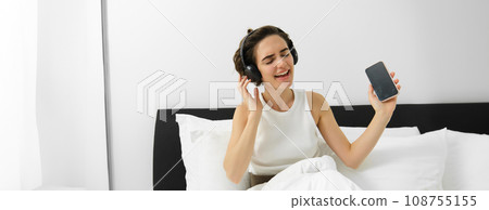 Portrait of happy woman waking up in bed, dancing while listening to music in wireless headphones, holding smartphone Portrait of happy woman waking up in bed, dancing while listening to music in wireless headphones, holding smartphone 108755155