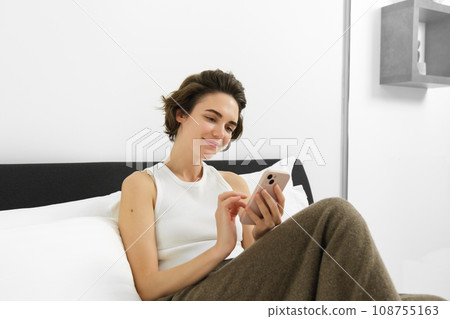 Portrait of young beautiful woman lying in bed, resting in bedroom, messaging, using mobile phone, holding smartphone and smiling, scrolling social media, spending comfort time at home Portrait of young beautiful woman lying in bed, resting in bedroom, messaging, using mobile phone, holding smartphone and smiling, scrolling social media, spending comfort time at home 108755163