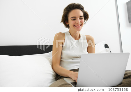 Image of smiling, happy young woman in bedroom, sitting on bed with laptop, laughing and looking at computer screen, working from home, freelancing Image of smiling, happy young woman in bedroom, sitting on bed with laptop, laughing and looking at computer screen, working from home, freelancing 108755168
