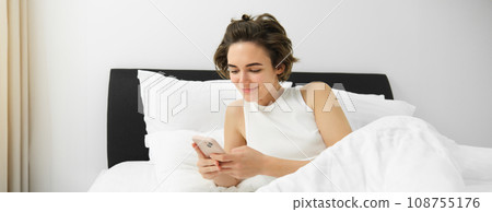 Portrait of young woman waking up in bed, using smartphone, turn off alarm on mobile phone, lying in her bedroom on white linen sheets 108755176