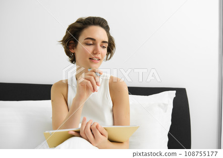 Portrait of young woman in bed, working or studying, writing in her journal, reading notebook notes, sharing thoughts with diary and smiling 108755177