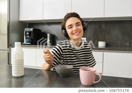 Portrait of happy smiling woman eating cereals with milk, drinking tea and listening music in wireless headphones, enjoys her morning, having breakfast alone in the kitchen 108755178