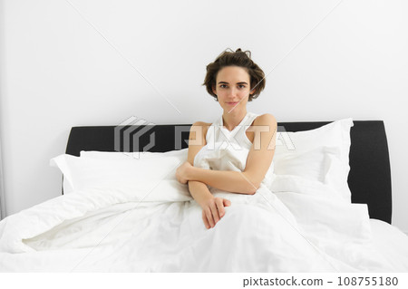 Image of young sensual woman with short hair, wearing tank top pyjama, lying in bed under white linen sheets, looking at camera, waking up in morning 108755180