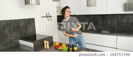 Happy woman dancing and cooking in the kitchen, listening music in earphones, making a meal, preparing salad, chopping vegetables on a counter 108755196
