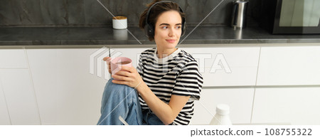 Portrait of stylish modern woman in headphones, listens music and drinks tea, eats cereals with milk for lunch, sits in kitchen, looks outside window with relaxed smile 108755322