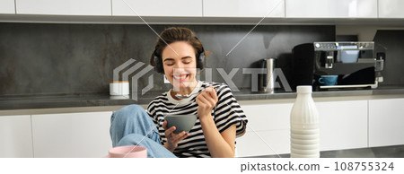 Portrait of smiling young candid woman, student eating morning cereals, having her breakfast, listening music in wireless headphones, sitting in the kitchen 108755324
