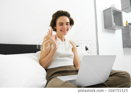 Image of young woman working from home, connects to online video chat, team meeting, waving hand at laptop, sitting on bed, studying, e-learning from home Image of young woman working from home, connects to online video chat, team meeting, waving hand at laptop, sitting on bed, studying, e-learning from home 108755325