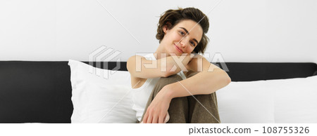 Close up portrait of young female model in white tank-top, sitting on bed in her bedroom and smiling at camera, leaning on pillows 108755326