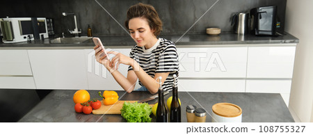 Portrait of young woman with smartphone, sitting in the kitchen and cooking salad, searching for healthy recipe online, has vegetables and chopping board on the table Portrait of young woman with smartphone, sitting in the kitchen and cooking salad, searching for healthy recipe online, has vegetables and chopping board on the table 108755327