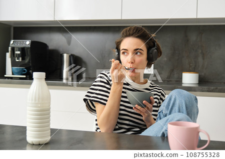Cute modern woman, student eating quick breakfast, having cereals with milk and coffee, listening to podcast or music in wireless headphones Cute modern woman, student eating quick breakfast, having cereals with milk and coffee, listening to podcast or music in wireless headphones 108755328