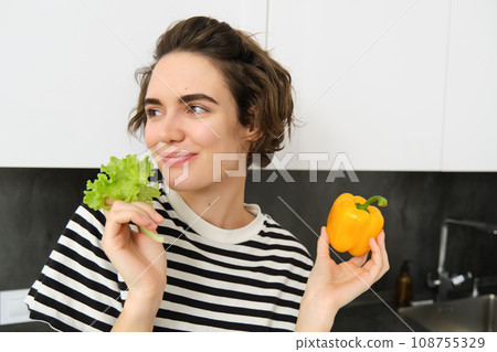 Portrait of smiling brunette woman, holding lettuce leaf and yellow sweet pepper, eating vegetables, likes fresh vegies, healthy diet concept Portrait of smiling brunette woman, holding lettuce leaf and yellow sweet pepper, eating vegetables, likes fresh vegies, healthy diet concept 108755329