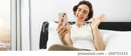 Friendly smiling woman talking with friend on smartphone app, video call someone while resting in bed, lying in bedroom 108755335