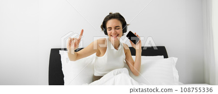 Image of beautiful smiling woman, dancing on bed, waking up and listening to music in wireless headphones, energizing, happy morning 108755336