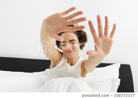 Close up portrait of carefree, beautiful young woman stretching her arms and smiling with dreamy, closed eyes, waking up in morning after good night sleep 108755337