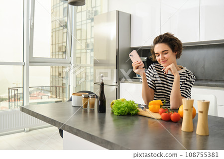 Portrait of smiling beautiful woman in the kitchen, looking at vegetables and holding smartphone, deciding what to cook, searching for ideas for lunch meals 108755381