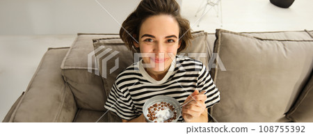 Close up of cute young female model, eating cereals with milk, enjoys her breakfast on sofa in living room, smiling and looking happy Close up of cute young female model, eating cereals with milk, enjoys her breakfast on sofa in living room, smiling and looking happy 108755392