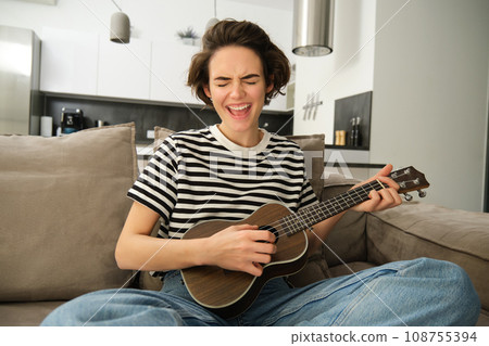Beautiful girl playing ukulele on sofa, singing her favourite song and picking chords on small guitar, sitting in living room Beautiful girl playing ukulele on sofa, singing her favourite song and picking chords on small guitar, sitting in living room 108755394