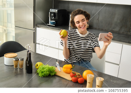 Smiling young woman, food blogger recording a video on smartphone with food, cooking and making selfie, taking pictures during meal preparation, sitting near vegetables and holding an apple Smiling young woman, food blogger recording a video on smartphone with food, cooking and making selfie, taking pictures during meal preparation, sitting near vegetables and holding an apple 108755395