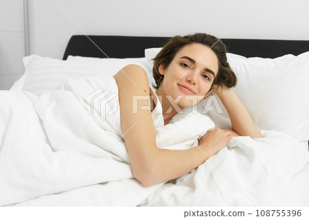Image of young woman lying in bed, resting at home under white linen sheets, looking with pleased smile, waking up and relaxing Image of young woman lying in bed, resting at home under white linen sheets, looking with pleased smile, waking up and relaxing 108755396