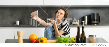 Cute young woman, lifestyle blogger cooking, sitting in the kitchen with vegetables and chopping board, taking selfie on her new smartphone, sending kisses at mobile camera 108755427
