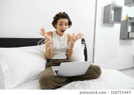 Image of woman sitting on bed, looking at smartphone with surprised, shocked face expression, has amazed reaction to online deal 108755447