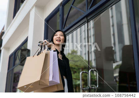 Young asian woman in shopping. Fashion woman in black with shopping bag walking around the city after shopping. Black friday Young asian woman in shopping. Fashion woman in black with shopping bag walking around the city after shopping. Black friday 108756442