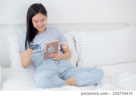 Young asian woman sitting on bed using digital tablet shopping online with credit card. 108758435