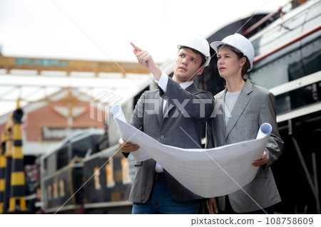 Young caucasian engineer man and woman in suit checking train looking blueprint in station. Young caucasian engineer man and woman in suit checking train looking blueprint in station. 108758609