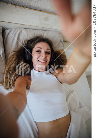 Woman taking pictures of herself in pajamas in bedroom on bed 108764337