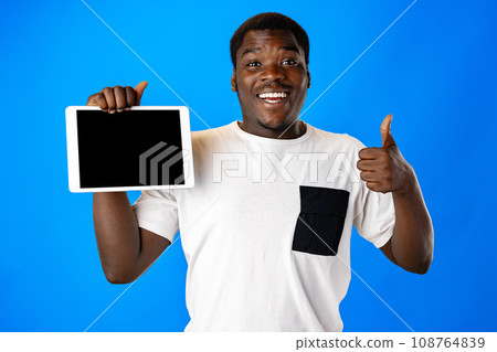 Young african man showing digital tablet with black screen against blue background Young african man showing digital tablet with black screen against blue background 108764839