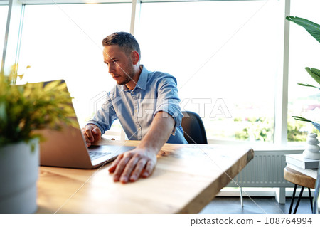 Portrait of young man sitting at his desk in the office and using computer Portrait of young man sitting at his desk in the office and using computer 108764994