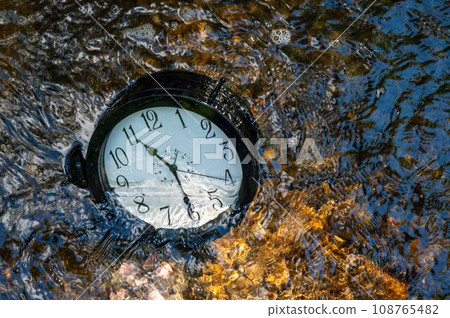 A large clock half submerged in a flowing stream with ripples pebbles A large clock half submerged in a flowing stream with ripples pebbles 108765482