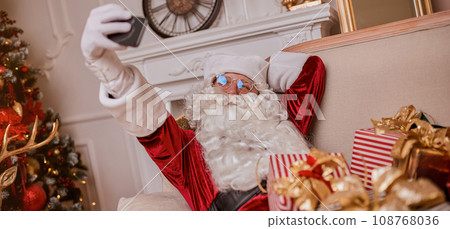 Santa Claus making selfie on phone sitting with christmas gifts. New year and Merry Christmas Santa Claus making selfie on phone sitting with christmas gifts. New year and Merry Christmas 108768036