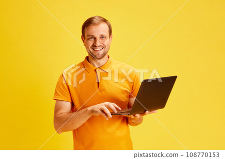 Portrait of young attractive man, smart guy, student holding laptop and looking at camera while working or studying isolated yellow studio background. 108770153
