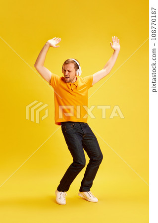 Full-length portrait of young man wearing style casual outfit and dancing listening music in headphones isolated yellow studio background. 108770187