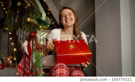 Happy cheerful woman in pajamas sitting on wooden stairs and holding Christmas present in gift box she received on winter holidays 108770321