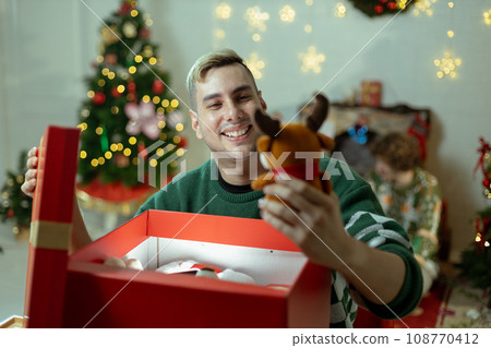 Christmas day concept. handsome man sitting surprised and opening gift box with christmas tree Christmas day concept. handsome man sitting surprised and opening gift box with christmas tree 108770412